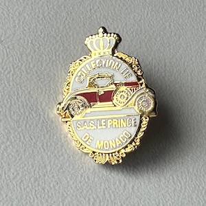 Könnte beinhalten: Goldfarbene Anstecknadel mit einem weißen und roten Autodesign, mit dem Text "COLLECTION DE SAS LE PRINCE DE MONACO". Die Anstecknadel hat eine Krone oben und einen verzierten Rand.