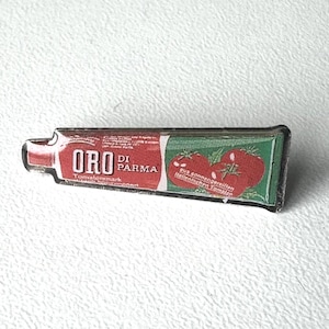 Könnte beinhalten: Ein rechteckiger Ansteckpin mit einem Design, das an eine Tube Tomatenmark erinnert. Der Pin hat ein rot-grünes Farbschema mit dem Text "ORO di Parma" und einer Illustration von Tomaten.