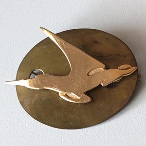 Pode incluir: Broche vintage em metal dourado com um pássaro estilizado em voo, sobre um fundo circular de latão envelhecido. As asas do pássaro estão estendidas, criando um design dinâmico. Um pequeno orifício é visível no círculo de latão.