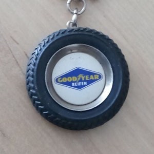 Könnte beinhalten: Ein Goodyear-Reifen-Schlüsselanhänger mit einem silbernen Schlüsselring. Der Schlüsselanhänger hat ein schwarzes Reifendesign mit einer silbernen Radkappe. Die Radkappe hat ein blau-weißes Diamantlogo mit den Worten "GOOD YEAR REIFEN".