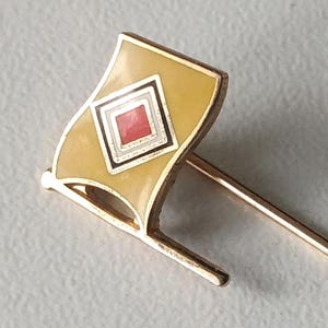 Op de afbeelding: Een goudkleurige vlaggenpin met een gele emaille vlag met een rood-wit ruitpatroon. De pin heeft een lange, slanke naald voor bevestiging, geschikt voor revers of hoeden.