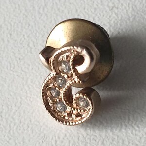 Könnte beinhalten: Ein goldfarbener Ohrstecker mit dem Buchstaben "E" und kleinen, klaren Edelsteinen. Der Ohrring hat einen Steckerverschluss und ist vor einem weißen Hintergrund platziert. Das Design ist verziert und detailliert.
