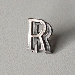 Könnte beinhalten: Eine silberfarbene Anstecknadel mit den ineinander verschlungenen Buchstaben "RR". Die Anstecknadel hat ein dreidimensionales Design mit leicht gealtertem Aussehen. Die Anstecknadel befindet sich vor einem hellgrauen Hintergrund.