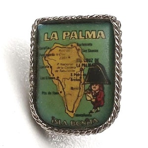 Peut inclure: Une épingle argentée avec une carte verte et jaune de La Palma, îles Canaries. L'épingle porte le texte "LA PALMA" et une petite illustration d'une personne tenant un parapluie.
