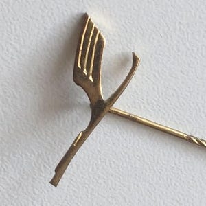 Pode incluir: Alfinete dourado com um design estilizado. O alfinete apresenta uma cabeça decorativa com uma forma geométrica e um longo pino pontiagudo. O alfinete está sobre um fundo branco.
