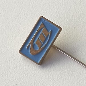 May include: A vintage lapel pin featuring a rectangular blue enamel face with a gold design. The pin has a long, silver needle. A second, smaller pin is also visible.