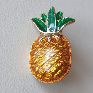 Op de afbeelding: Een goudkleurige ananasvormige speld met een getextureerd oppervlak. De ananas heeft een geel-oranje lichaam en groene bladeren aan de bovenkant. De speld is waarschijnlijk gemaakt van metaal en heeft een glanzende afwerking.