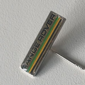 Könnte beinhalten: Eine silberne Anstecknadel mit einem schwarzen, grünen und gelben gestreiften Rechteck mit dem Text "RANGE ROVER" in Schwarz.