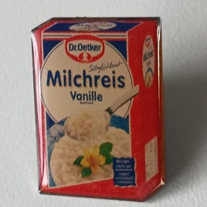 Può includere: Una piccola scatola rettangolare di Dr. Oetker Milchreis Vanille. La scatola è rossa, bianca e blu con il nome del prodotto e un'immagine del dessert. L'immagine mostra un primo piano della scatola su una superficie bianca.