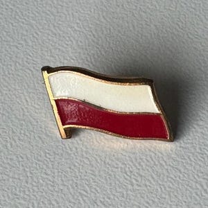 Puede incluir: Un pequeño pin dorado con la bandera polaca. El diseño de la bandera tiene una franja blanca sobre una franja roja. El pin tiene un borde dorado y está sobre una superficie gris claro texturizada.