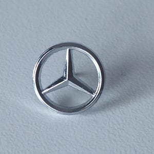 May include: A silver Mercedes-Benz emblem, a three-pointed star inside a circle. The emblem is displayed against a light grey background, highlighting its polished surface and iconic design. The emblem is a symbol of the Mercedes-Benz brand.