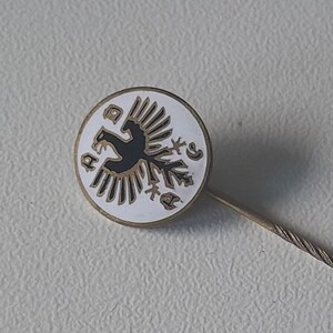 May include: A white enamel pin with a black eagle design and the letters "ADC" around the edge. The pin has a gold-coloured metal pin back.