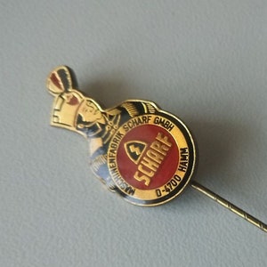 Könnte beinhalten: Goldfarbene Metallsticknadel mit einem rot-schwarz-weißen Logo, auf dem "Maschinenfabrik Scharf GmbH" und "SCHARF" mit dem Text "0-4700 HAMM" steht. Die Nadel ist an einer dünnen, goldfarbenen Metallstift befestigt.