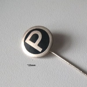 Op de afbeelding: Een zilveren emaille-speld met een zwarte cirkel en een witte letter "P" in het midden. De speld is ongeveer 15 mm in diameter en heeft een lange, dunne speld aan de achterkant. De tekst "Hoffstätter Bonn" is zichtbaar op de achterkant van de speld.