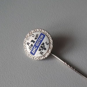 May include: Silver enamel pin with blue and white lettering that reads "AMC IDSTEIN" surrounded by a silver laurel wreath. The pin is attached to a silver pin back. The pin is 14 mm in diameter.