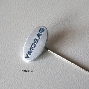 Könnte beinhalten: Silberne Anstecknadel mit einem runden, ovalen Kopf. Der Kopf trägt den Text "YMOS AG". Die Nadel ist etwa 15 mm x 8 mm groß.