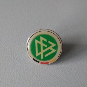 Puede incluir: Un pin verde, blanco y negro con el logotipo de la Federación Alemana de Fútbol. El pin es redondo y tiene un respaldo plateado. El texto "DFB - Deutscher Fußball Bund Pin frühe 2000er" y "DFB - German Football Association Pin early 2000s" está debajo del pin.