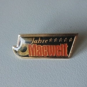 May include: A gold-coloured pin with a black and orange design. The pin reads "5 Jahre MACWELT Pin von 2002". The pin is 2.7 cm by 1 cm.