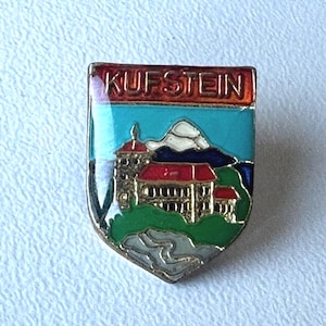 Könnte beinhalten: Ein schildförmiger Emaille-Pin mit dem Wort "KUFSTEIN" oben. Der Pin zeigt ein Schloss mit rotem Dach, Berge und blauen Himmel. Der Pin hat einen silberfarbenen Rand.
