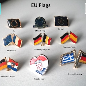 Può includere: Una collezione di spille smaltate con bandiere di paesi europei. Le spille sono disposte in file e includono bandiere di Germania, Spagna, Francia, Belgio, Croazia, Grecia, Italia e Unione Europea. Le spille sono tutte dorate e hanno una finitura lucida.