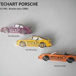 May include: Three enamel pins featuring Porsche cars from the 1990s. The pins are made by TechArt and feature a 911 993 in purple, a 911 993 in yellow, and a Boxster in orange.