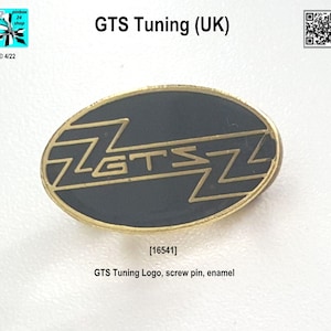 以下が含まれることがあります： スタイリッシュなフォントで「GTS」という文字が書かれた黒と金のエナメルピン。ピンは楕円形で、ネジ式の留め具が付いています。