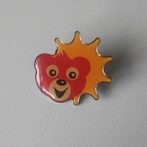 Puede incluir: Un pin de esmalte rojo y amarillo con un oso de dibujos animados con una cara sonriente y un sol detr&aacute;s.