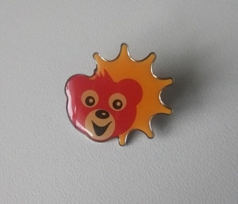 Puede incluir: Un pin de esmalte rojo y amarillo con un oso de dibujos animados con una cara sonriente y un sol detr&aacute;s.
