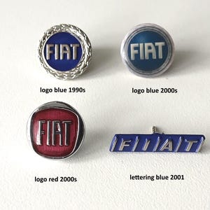 Op de afbeelding: Vier verschillende versies van het FIAT-logo, die de evolutie van het ontwerp in de loop van de tijd laten zien. De logo's zijn allemaal zilver en blauw, met uitzondering van het rode logo uit de jaren 2000.