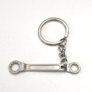 Puede incluir: Un llavero plateado con forma de llave inglesa. El llavero presenta un diseño de llave inglesa pequeño y detallado con enchufes abiertos en cada extremo. La llave está unida a una cadena y a un llavero redondo.