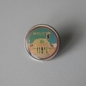 Puede incluir: Un pin redondo de color plateado con una imagen colorida de un edificio y el texto "BERLIN" en letras doradas. El edificio est&aacute; rodeado de una valla y tiene un techo verde. El pin tambi&eacute;n presenta un emblema de oso.