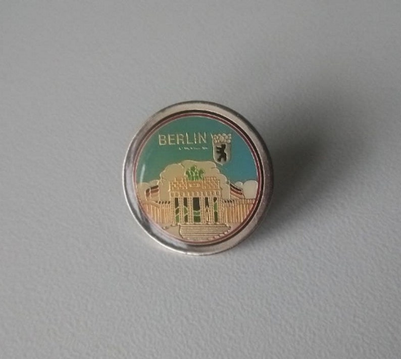 Puede incluir: Un pin redondo de color plateado con una imagen colorida de un edificio y el texto "BERLIN" en letras doradas. El edificio est&aacute; rodeado de una valla y tiene un techo verde. El pin tambi&eacute;n presenta un emblema de oso.