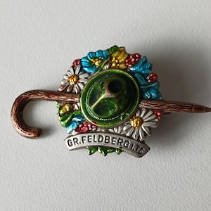 May include: A vintage brooch featuring a green and silver umbrella design, adorned with colorful flowers and the text "GR.FELDBERG I.TS". The brooch has a brown handle and a pointed end, with a detailed floral arrangement.