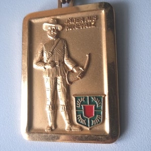 Könnte beinhalten: Ein goldfarbener Metallanhänger mit einem Reliefbild eines Mannes in Hut und Uniform, der ein Jagdhorn hält. Der Text "JAGER AUS KURPFALZ" ist über dem Mann eingraviert. Ein Schild mit roter Mitte und grünem Rand befindet sich unter dem Mann.