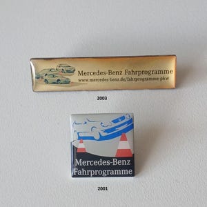 Puede incluir: Dos pines vintage de Mercedes-Benz Fahrprogramme. El pin rectangular de 2003 presenta un fondo dorado con ilustraciones de coches. El pin cuadrado de 2001 tiene un fondo plateado con un gráfico de coche y conos de tráfico.