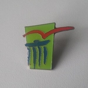 Puede incluir: Pin de esmalte verde, rojo y azul con un dise&ntilde;o abstracto. El pin presenta una l&iacute;nea roja que se curva sobre un dise&ntilde;o azul y verde.