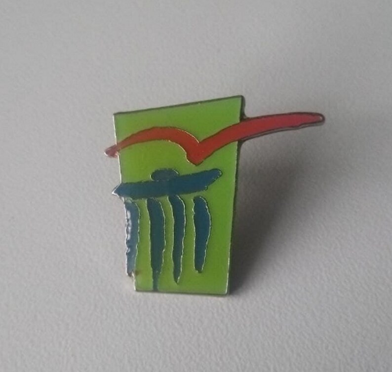 Puede incluir: Pin de esmalte verde, rojo y azul con un dise&ntilde;o abstracto. El pin presenta una l&iacute;nea roja que se curva sobre un dise&ntilde;o azul y verde.