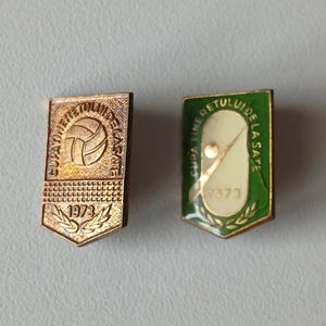 May include: Two vintage enamel pins. One is gold-toned with a volleyball design and the year 1973. The other is green and white with a stylized design and the year 1973. Both pins have text in Spanish.