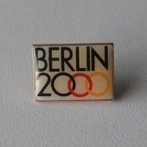 Puede incluir: Un pin blanco y dorado con el texto "BERLIN 2000" y cinco anillos entrelazados en rojo, amarillo y azul.