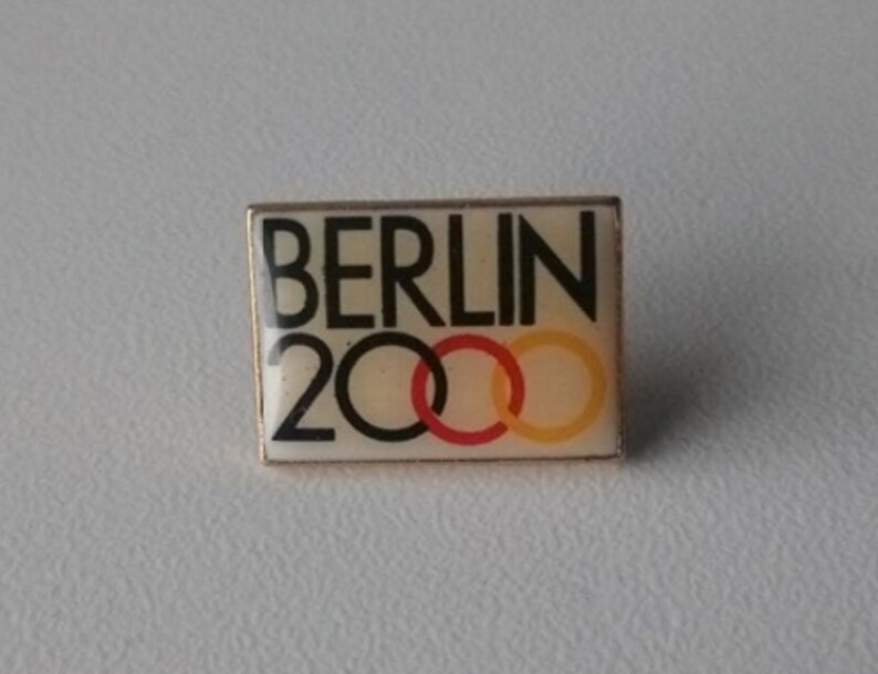 Puede incluir: Un pin blanco y dorado con el texto "BERLIN 2000" y cinco anillos entrelazados en rojo, amarillo y azul.