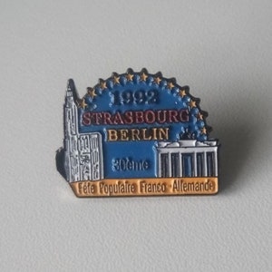 Puede incluir: Un pin de esmalte azul y dorado que representa la Catedral de Estrasburgo y la Puerta de Brandeburgo. El pin conmemora el Festival Popular Franco-Alem&aacute;n de 1992 e incluye el texto "1992 Strasbourg Berlin 30eme Fete Populaire Franco-Allemande".