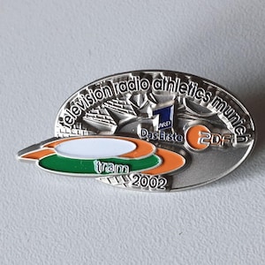 Könnte beinhalten: Silberne Anstecknadel mit der Aufschrift "television radio athletics munich" und den Logos von ARD und ZDF. Die Anstecknadel zeigt auch ein Straßenbahndesign mit der Aufschrift "tram 2002".