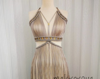 May include: A beige macrame dress with gold beads and fringe. The dress has a deep V-neckline and a cut-out design at the waist.