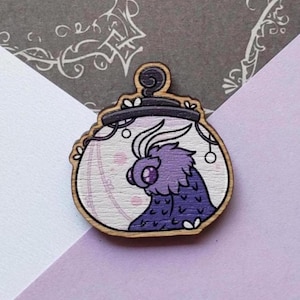 May include: Wooden pin depicting a purple creature within a decorative frame. The creature has large eyes and feathery details. The frame is outlined in black and has a small loop at the top. The background is a light purple with pink accents.