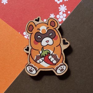 May include: Wooden bear-like character holding a red pepper. The bear has orange fur, a brown mask, and a white belly. The bear is on a background of orange, red, and brown paper.