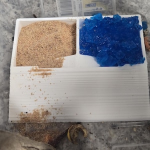 May include: A white plastic container divided into two sections. One section is filled with light brown, granular material, and the other with bright blue, crystalline pieces. The container sits on a gray surface.