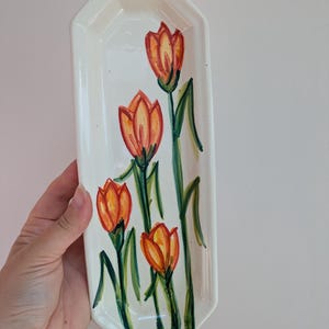 May include: A long, narrow, off-white ceramic tray with an octagonal shape. The surface features a hand-painted design of four orange and yellow tulips with green stems and leaves, arranged vertically along the centre of the elongated dish.