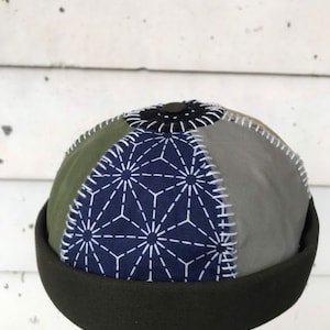 May include: A green, blue, and gray patchwork beanie with a geometric pattern and white stitching. The beanie has a dark green fold-over brim.