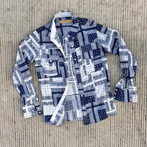 Può includere: Una camicia abbottonata a maniche lunghe con un design patchwork nei toni del blu e del bianco. La camicia presenta un colletto classico, polsini con bottoni e due tasche sul petto. Il tessuto ha un motivo simile al paisley.