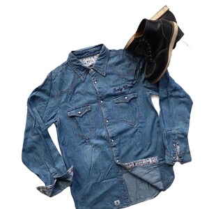 May include: A blue denim long-sleeve shirt with a button-down collar and a pocket on the left chest. The shirt has a white and black patterned lining on the inside of the cuffs. The shirt has the text "Body Glove" embroidered on the right chest pocket.